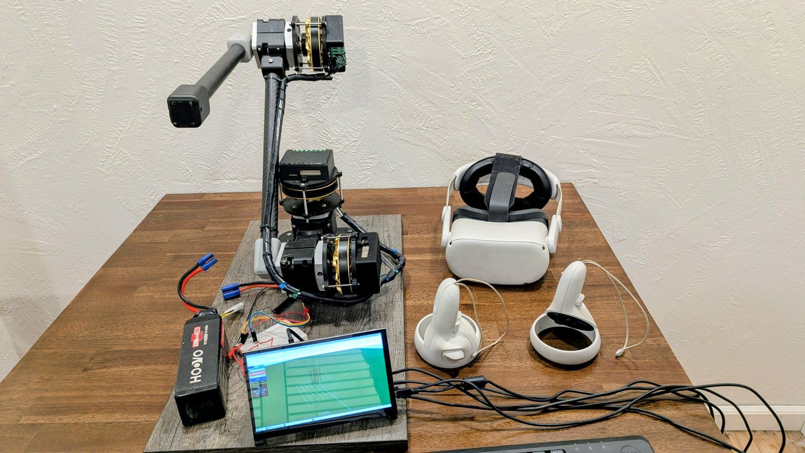 A multi-axis robotic arm with carbon-fiber links and brushless joint actuators on a workbench, beside a VR headset, two VR controllers and a control display.